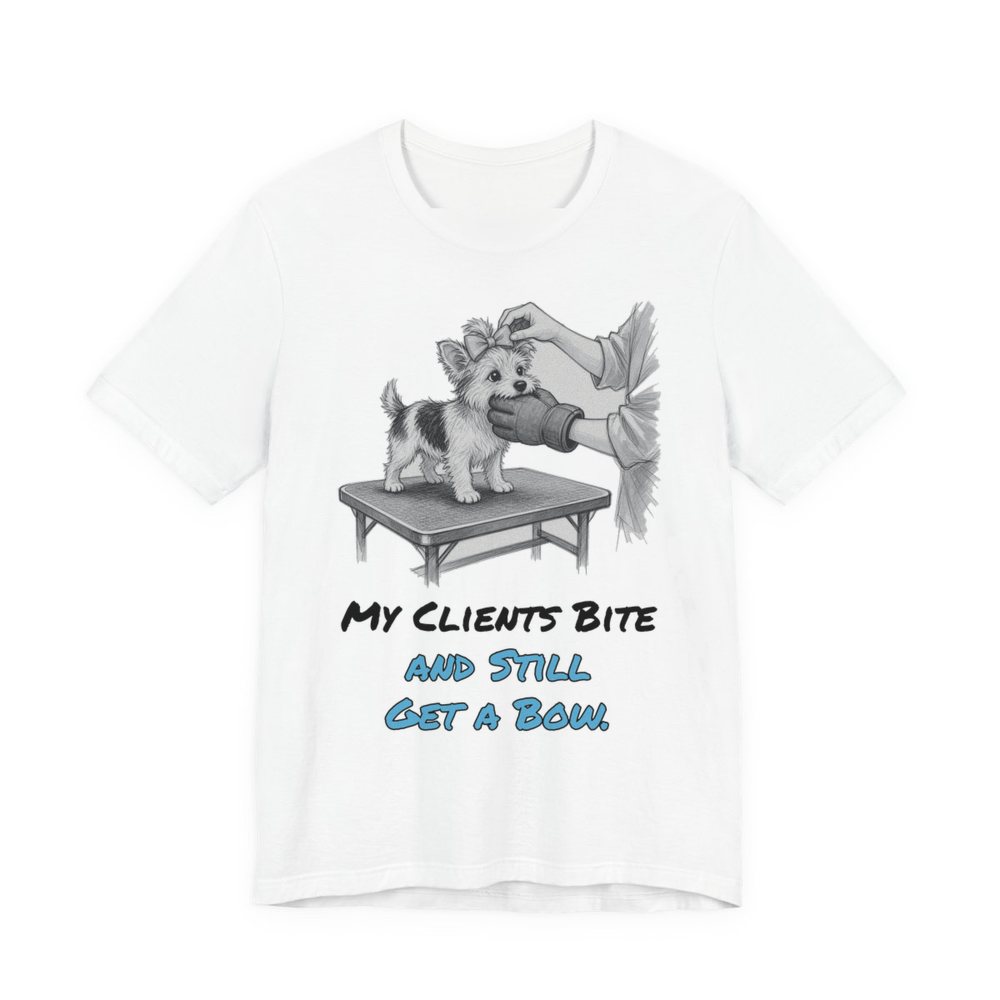 White t-shirt printed in full-color print with a full-color pencil drawing of a small dog getting a bow on a grooming table, nipping at the groomer's glove and the words “My Clients Bite and Still Get a Bow.”