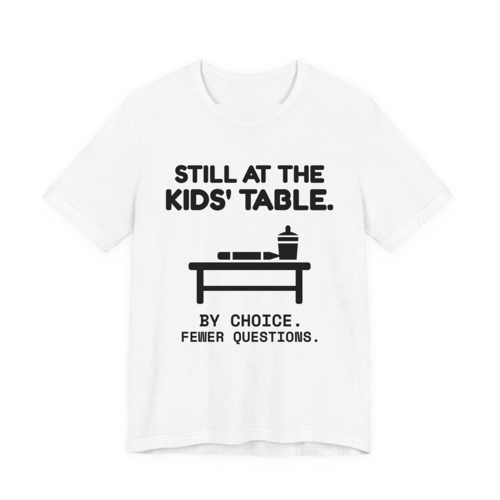 White t-shirt printed in dark ink with the words “Still at the Kids' Table.”