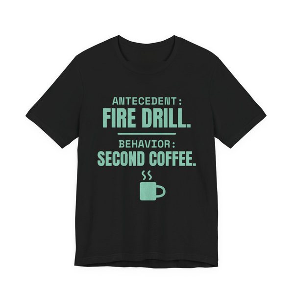 Black t-shirt printed in chalk mint ink with a steaming mug and the words “Antecedent: Fire Drill. Behavior: Second Coffee.”