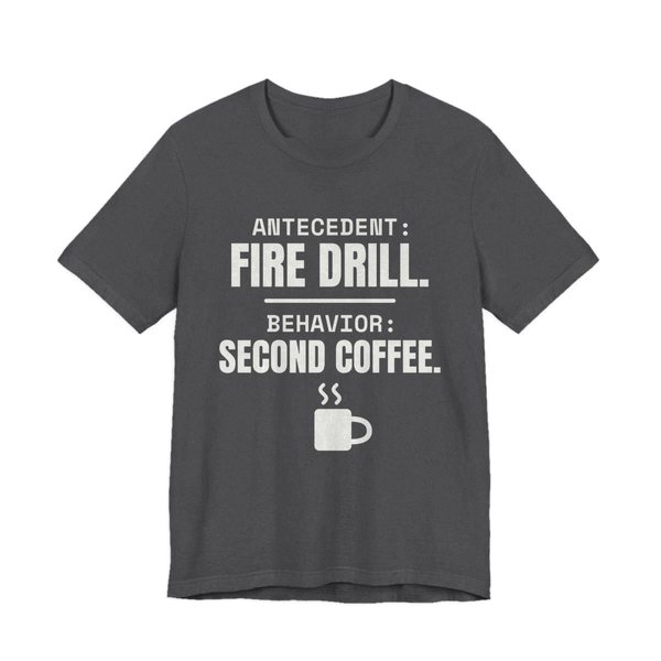 Dark Grey Heather t-shirt printed in off-white ink with a steaming mug and the words “Antecedent: Fire Drill. Behavior: Second Coffee.”