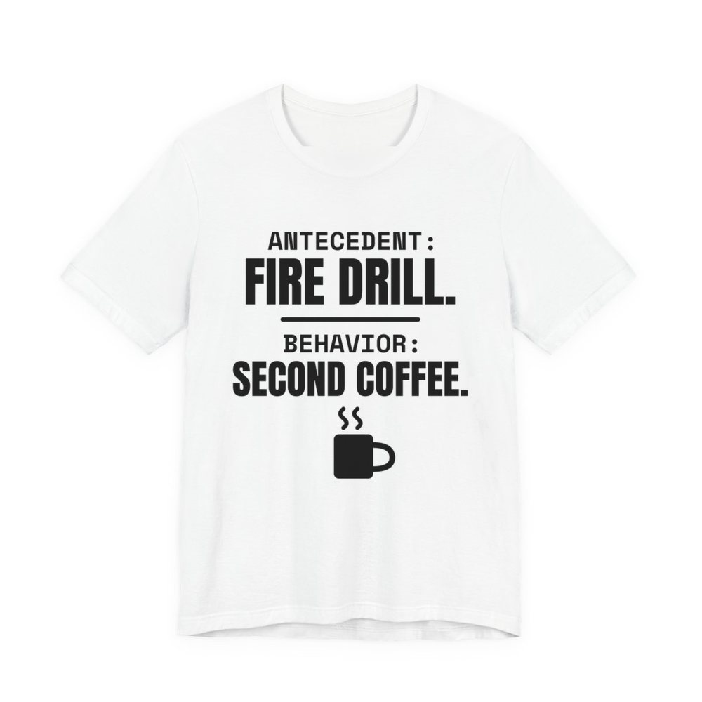 White t-shirt printed in dark ink with a steaming mug and the words “Antecedent: Fire Drill. Behavior: Second Coffee.”
