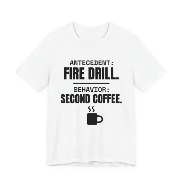 White t-shirt printed in dark ink with a steaming mug and the words “Antecedent: Fire Drill. Behavior: Second Coffee.”