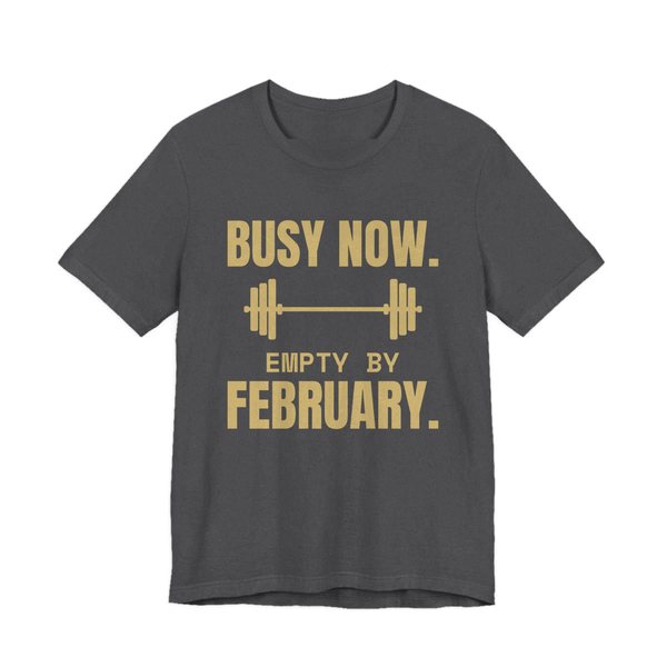 Dark Grey Heather t-shirt printed in champagne gold ink with a barbell and the words “Busy Now. Empty by February.”