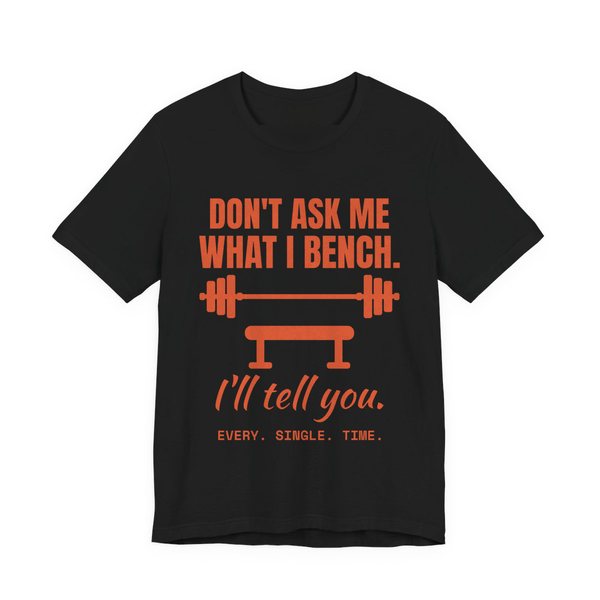 Black t-shirt printed in orange ink with a barbell over a bench and the words “Don't Ask Me What I Bench. I'll Tell You. Every. Single. Time.”