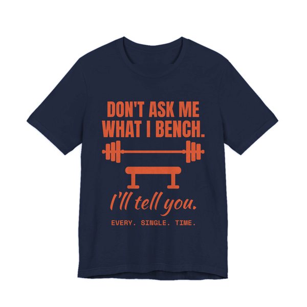 Navy t-shirt printed in orange ink with a barbell over a bench and the words “Don't Ask Me What I Bench. I'll Tell You. Every. Single. Time.”