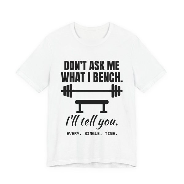 White t-shirt printed in dark ink with a barbell over a bench and the words “Don't Ask Me What I Bench. I'll Tell You. Every. Single. Time.”