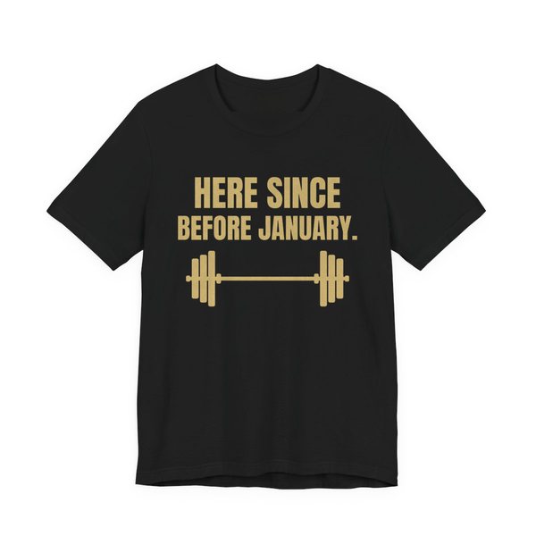 Black t-shirt printed in champagne gold ink with a barbell and the words “Here Since Before January.”