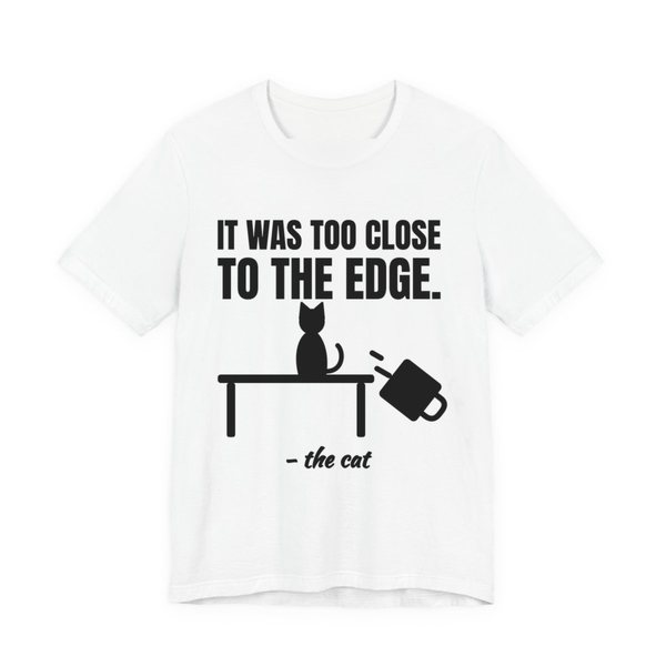 White t-shirt printed in dark ink with a cat on a table, a falling mug and the words “It Was Too Close to the Edge.”