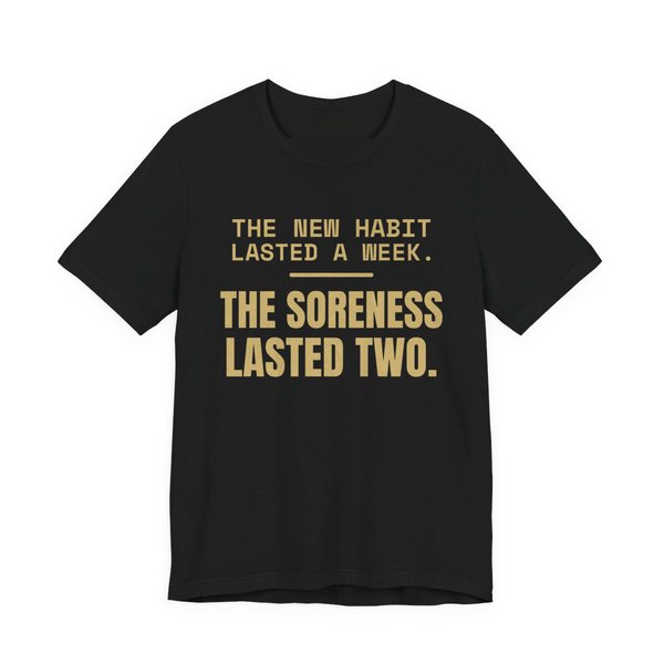 Black t-shirt printed in champagne gold ink with the words “New Habit Lasted a Week. The Soreness Lasted Two.”