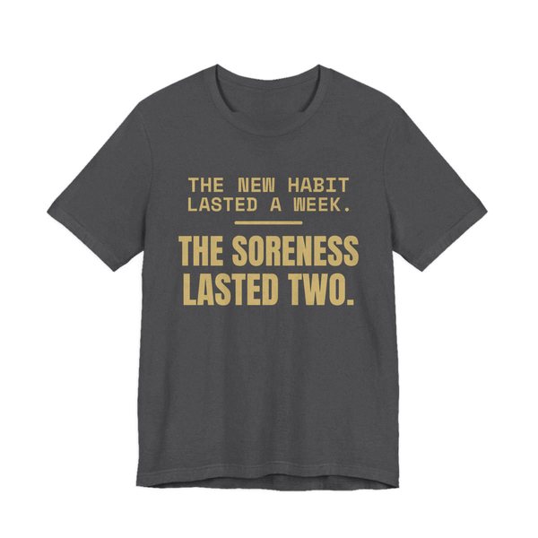 Dark Grey Heather t-shirt printed in champagne gold ink with the words “New Habit Lasted a Week. The Soreness Lasted Two.”