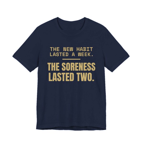 Navy t-shirt printed in champagne gold ink with the words “New Habit Lasted a Week. The Soreness Lasted Two.”