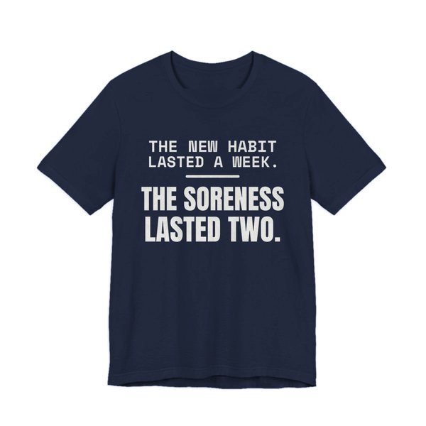 Navy t-shirt printed in off-white ink with the words “New Habit Lasted a Week. The Soreness Lasted Two.”